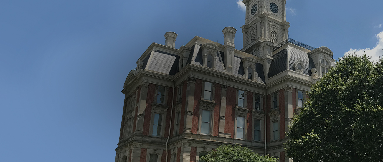 A grand historic building with red brick and stone accents, featuring a tall clock tower and mansard roofs, surrounded by trees and greenery.